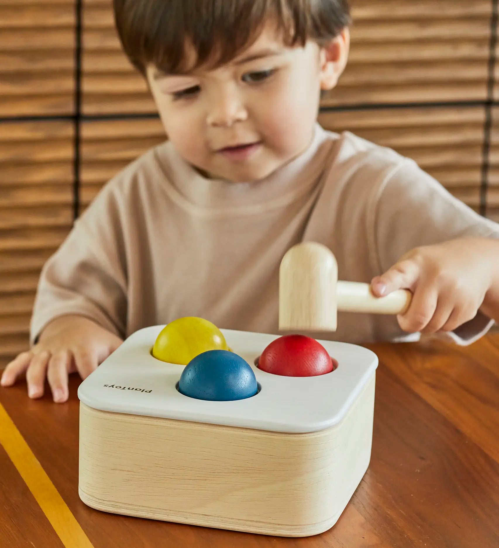 Toddler playing with the PlanToys punch and drop bin wooden pounding toy. Hammering the red ball into the bin through a slot on the lid.