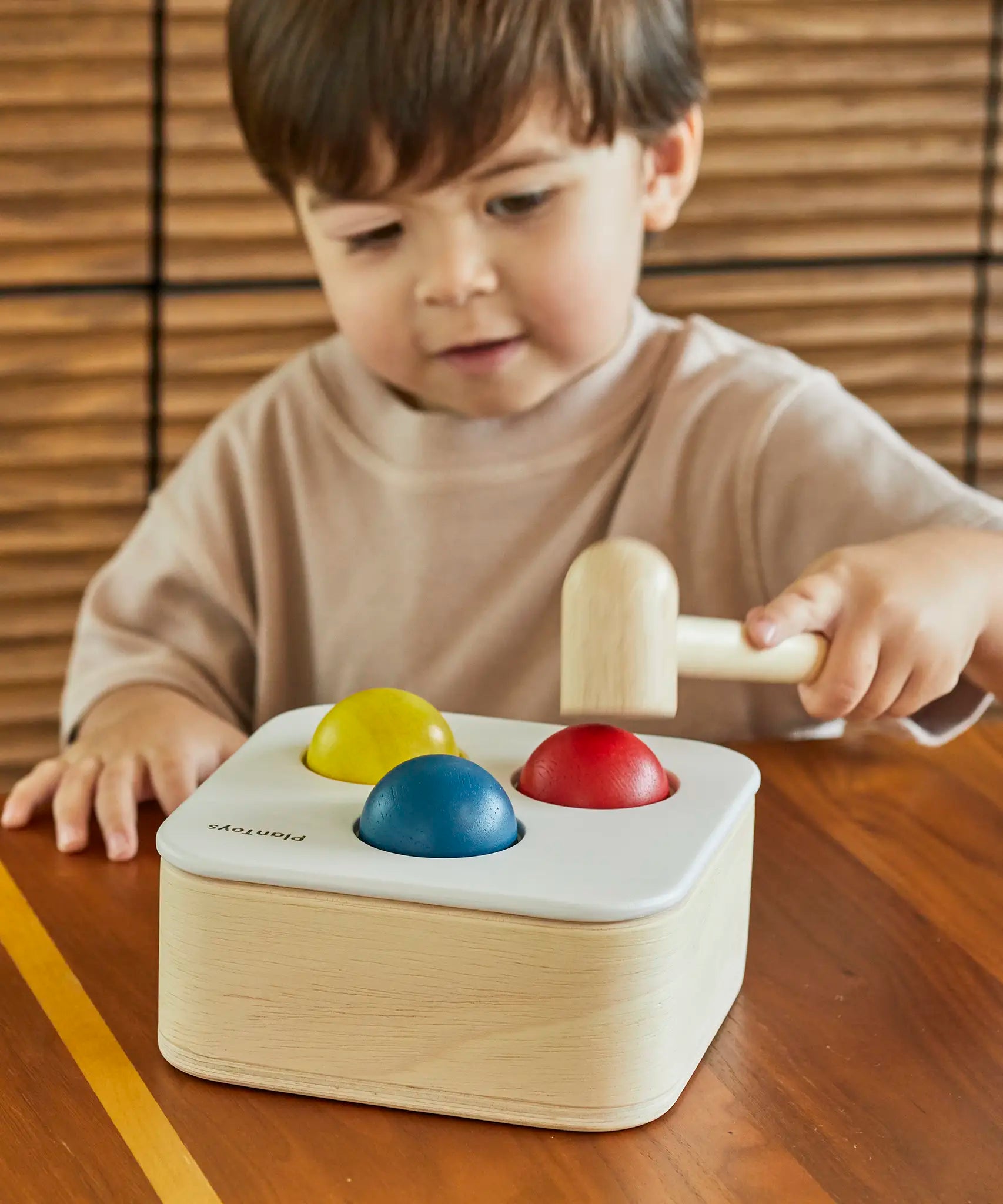 Toddler playing with the PlanToys punch and drop bin wooden pounding toy. Hammering the red ball into the bin through a slot on the lid.