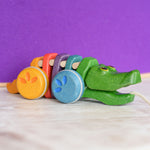 A Plan Toys Rainbow Alligator placed on a wooden surface in front of a purple wall. 