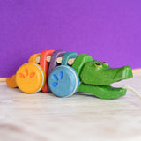 A Plan Toys Rainbow Alligator placed on a wooden surface in front of a purple wall. 