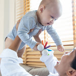 An adult holding up a baby, the baby has a PlanToys Baby Car Rattle in their hand. 