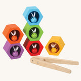 The PlanToys Bee Hives in the Rainbow colour way on a plain background. The bees are all in their matching coloured bee hive. The wooden tweezers can be seen placed in front of the hives. 
