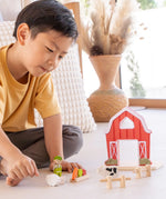 Child playing with the PlanToys barn house small world play set. Red wooden barn, mini veg patch, fences, cow, sheep, hen and farmer figure. 