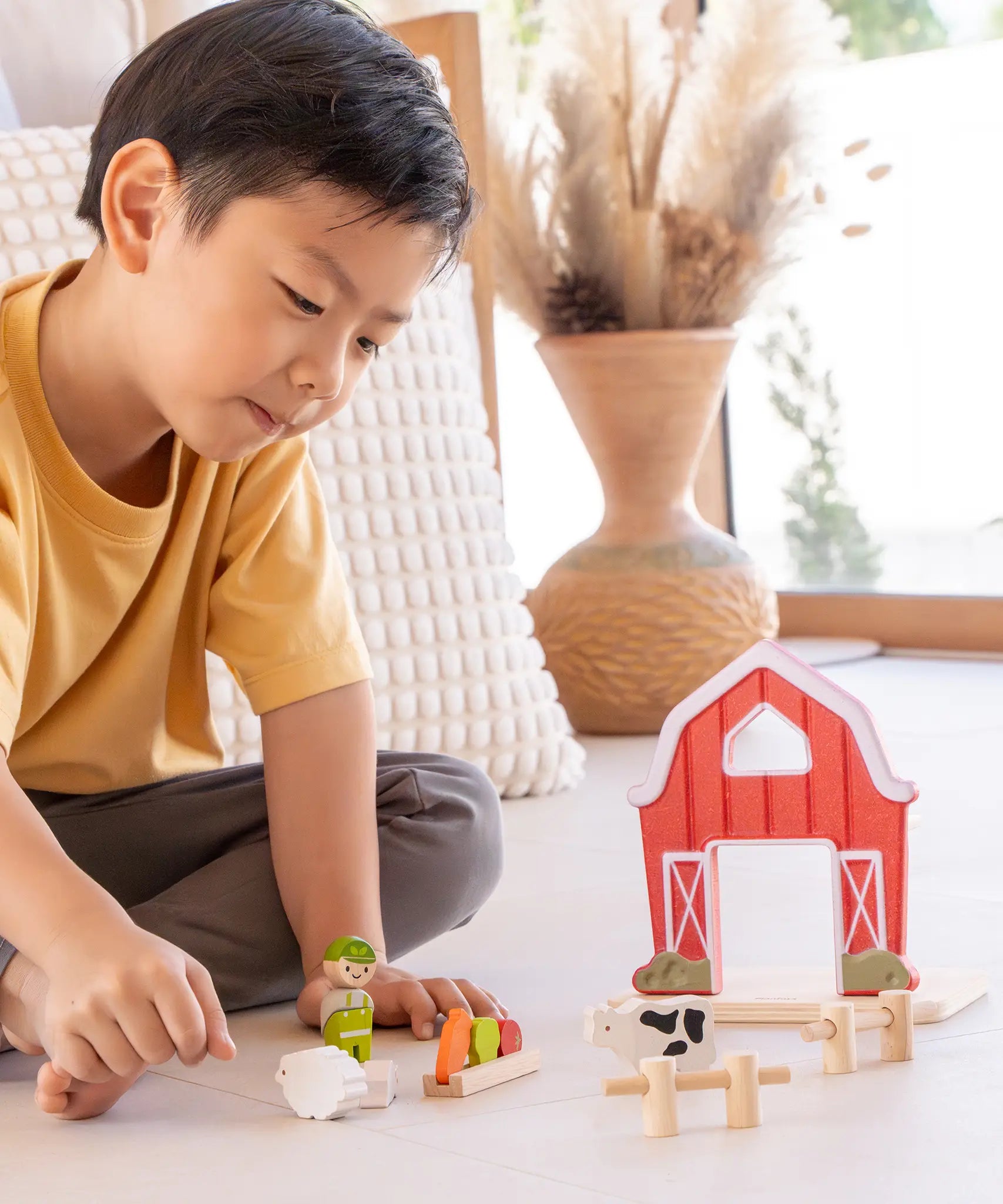 Child playing with the PlanToys barn house small world play set. Red wooden barn, mini veg patch, fences, cow, sheep, hen and farmer figure. 