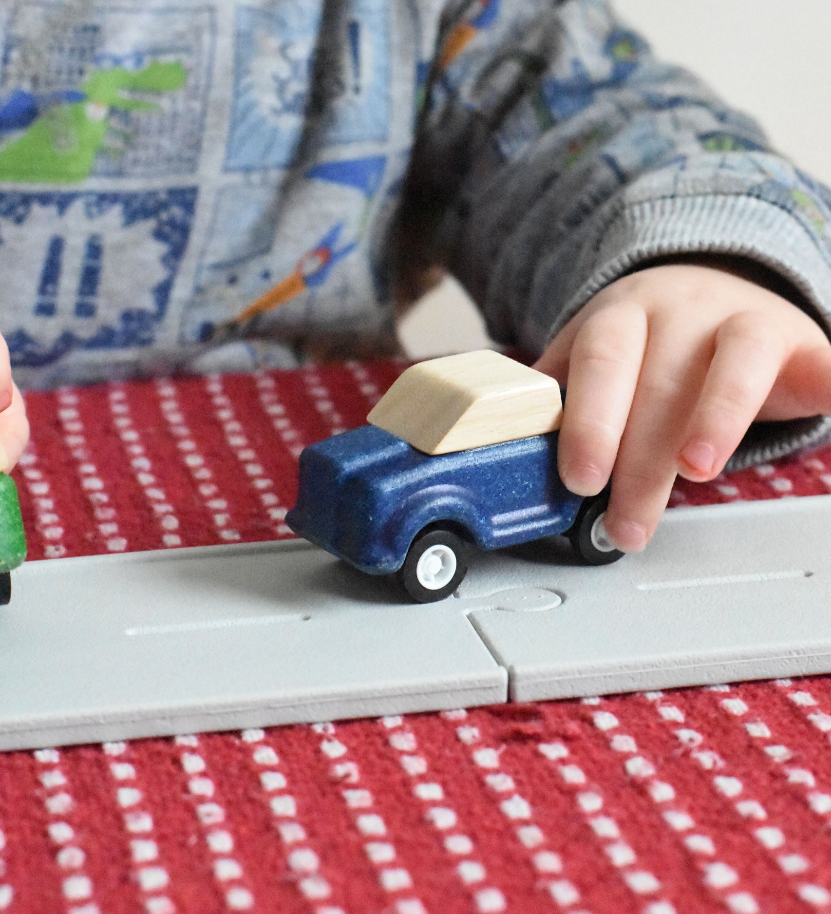A close up of a blue care on the PlanToys rubber road. A child's hand can be seen pushing the wooden toy car along the road.