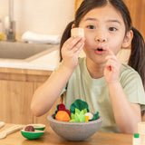 Child sitting at the table, holding a wooden PlanToys salt shaker with a bowl full of play salad foods