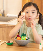 Child sitting at the table, holding a wooden PlanToys salt shaker with a bowl full of play salad foods