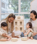 Two toddlers playing with the PlanToys wooden sensory drum musical toy with their parents striking the drums with the wood drumsticks.