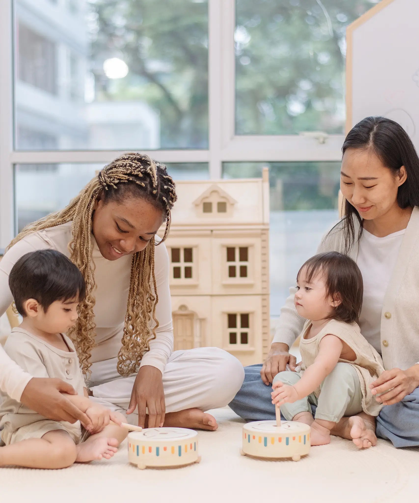 Two toddlers playing with the PlanToys wooden sensory drum musical toy with their parents striking the drums with the wood drumsticks.
