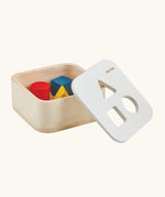 PlanToys shape and sort it out bin wooden shape sorter toy shown with the lid resting on the side. Square box with lid with three shaped holes in the lid. Yellow triangle, red cylinder and blue cube blocks.