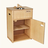 PlanToys wooden play sink kitchen unit. The counter top featuers a metal bowl sink and taps, a cut out handle door with a slotted shelf inside. 