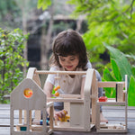 A child sitting on wooden decking outdoors playing with the PlanToys Slide N Go Dolls' House. 
