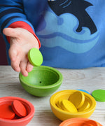 A close up of a child's hand playing with the Plan Toys Sort & Count Cups. The child has a green counter in their hand placing it into the green bowl.