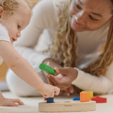 Child playing with the PlanToys sorting boat wooden shape sorting toy with an adult handing them a green cylinder shaped block. 