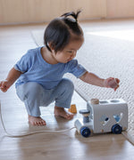 A toddler playing with the PlanToys orchard colourway Sorting Bus Baby Toy. They are kneeling down and have the cylinder shaped pieces in their hand. 