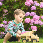 A child playing with the  Plan Toys Special Edition Natural Dump Truck outdoors on grass. There are purple hydrangeas behind them 