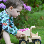 A child playing with the  Plan Toys Special Edition Natural Dump Truck outdoors on grass. There are purple hydrangeas behind them. The child is pushing the dumper truck along with hydrangea petals in the back of the truck. 