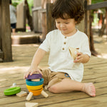 A child sitting on wooden decking outdoors playing with the PlanToys Stacking Rocket toy. 
