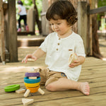A child sitting on wooden decking outdoors playing with the PlanToys Stacking Rocket toy. 
