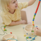 Child holding a lace filled with beads from the PlanToys threading beads wooden lacing toy set. The wooden storage box is in front of them.