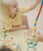 Child holding a lace filled with beads from the PlanToys threading beads wooden lacing toy set. The wooden storage box is in front of them.