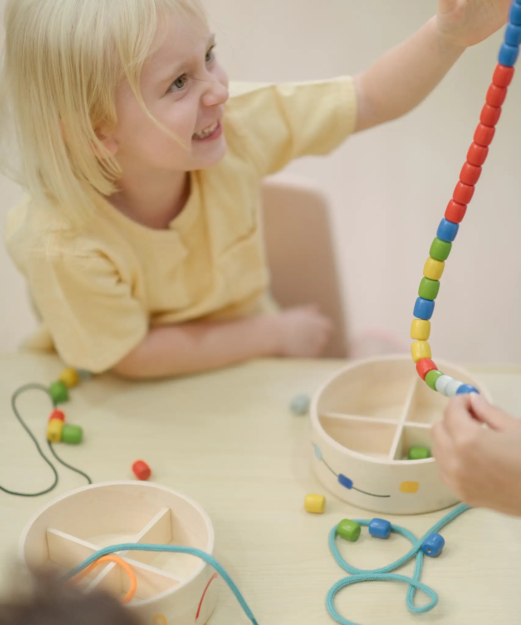 Child holding a lace filled with beads from the PlanToys threading beads wooden lacing toy set. The wooden storage box is in front of them.