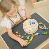 Child playing with the PlanToys threading beads wooden lacing toy. Rainbow coloured wooden beads with black and white laces in a handy wooden storage box with compartments. The set has been placed on the Plan Toys children's desk.