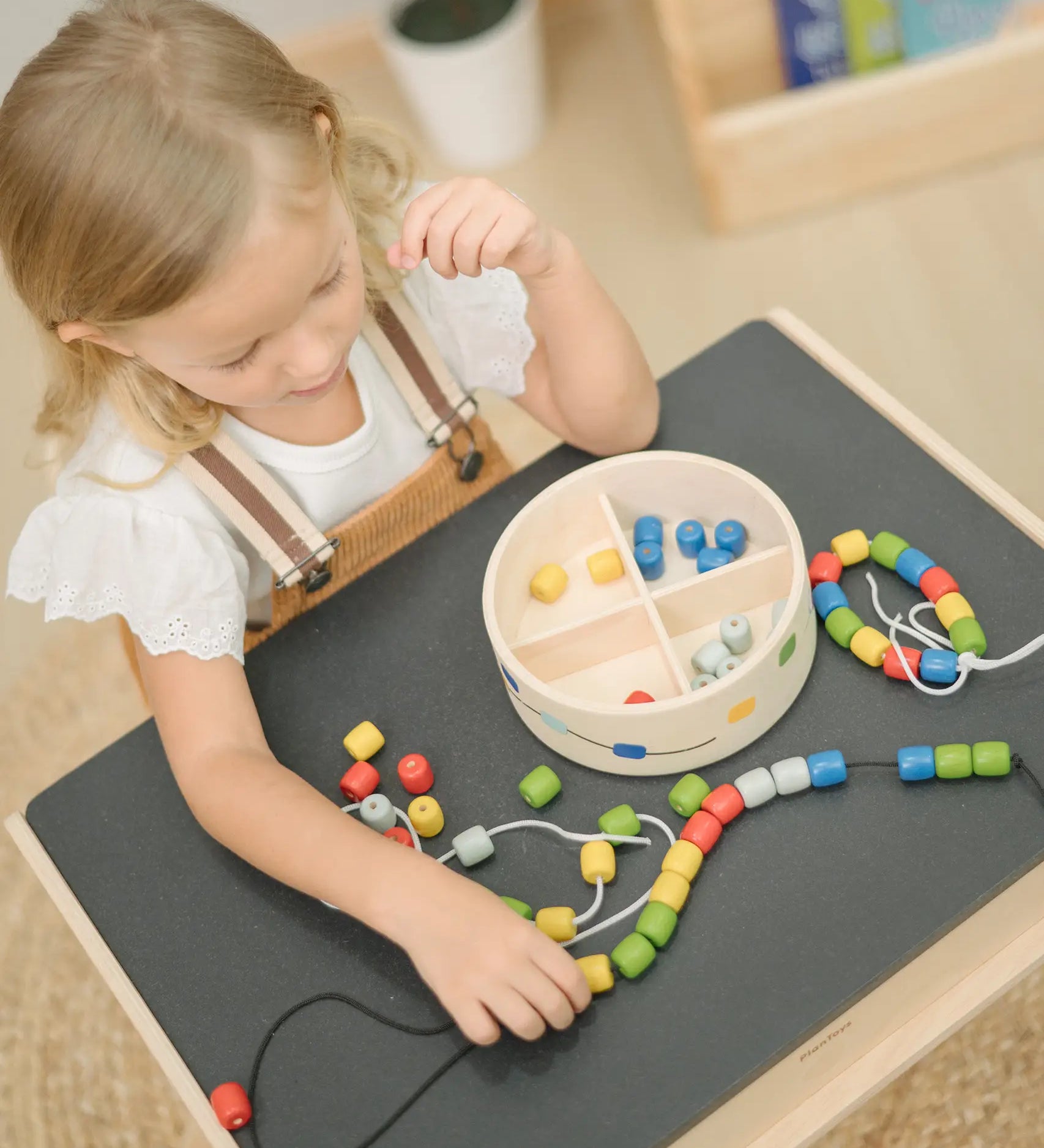 Child playing with the PlanToys threading beads wooden lacing toy. Rainbow coloured wooden beads with black and white laces in a handy wooden storage box with compartments. The set has been placed on the Plan Toys children's desk.