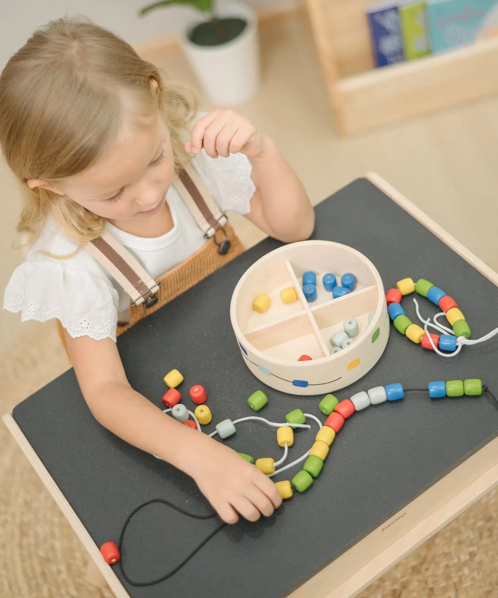 Child playing with the PlanToys threading beads wooden lacing toy. Rainbow coloured wooden beads with black and white laces in a handy wooden storage box with compartments. The set has been placed on the Plan Toys children's desk.