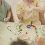 Children playing with the PlanToys threading beads wooden lacing toy, threading a grey coloured bead onto the white lace. Rainbow coloured wooden beads with black and white laces in a handy wooden storage box with compartments.