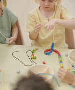 Children playing with the PlanToys threading beads wooden lacing toy, threading a grey coloured bead onto the white lace. Rainbow coloured wooden beads with black and white laces in a handy wooden storage box with compartments.