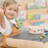 Child holding a lace filled with beads from the PlanToys threading beads wooden lacing toy set up to their neck as a necklace. The wooden storage box is in front of them on a PlanToys child's desk. 