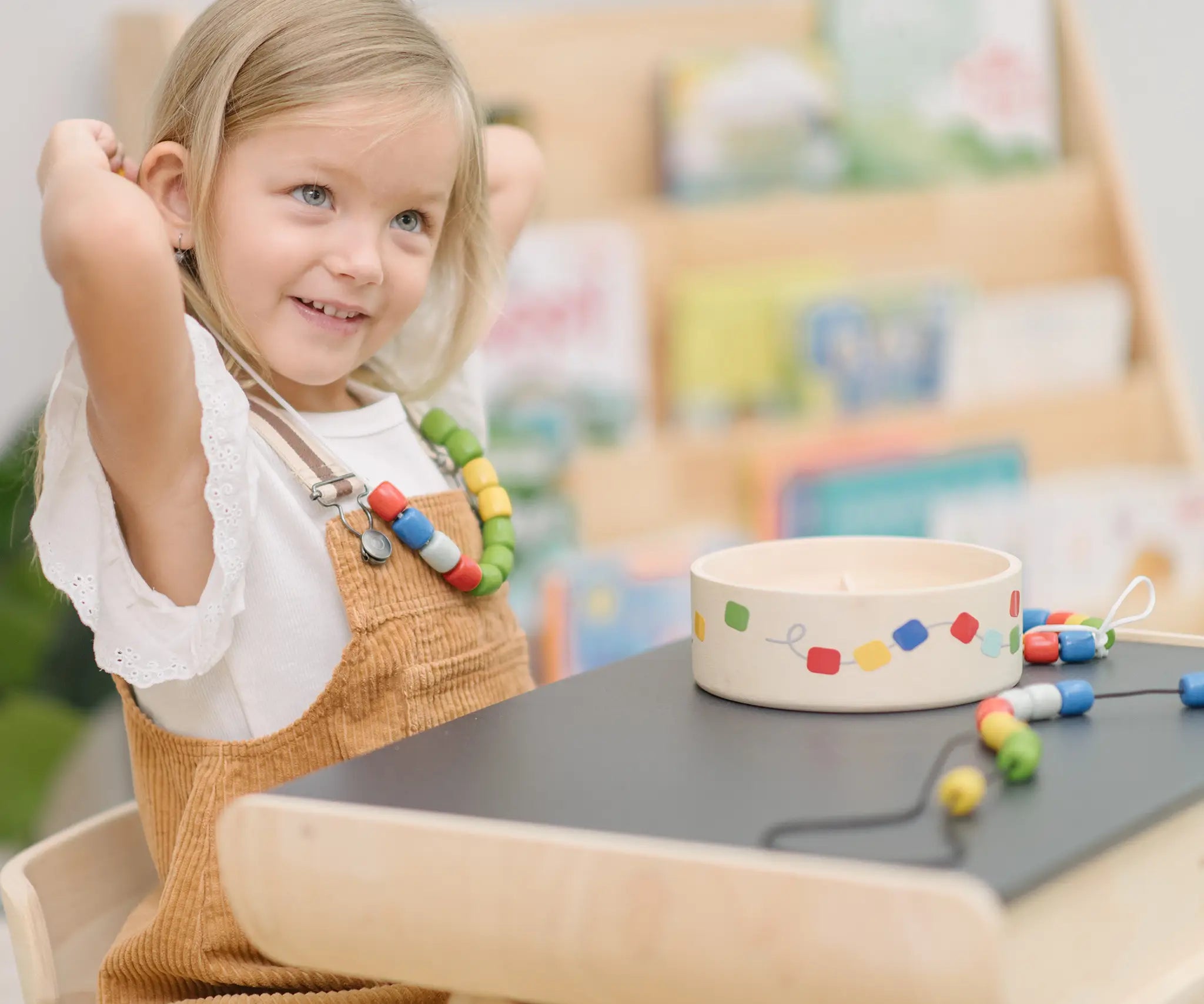 Child holding a lace filled with beads from the PlanToys threading beads wooden lacing toy set up to their neck as a necklace. The wooden storage box is in front of them on a PlanToys child's desk. 