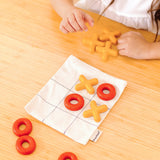 Child playing with the PlanToys Travel Tic-Tac-Toe wooden game set. Cotton bag printed with a grid with 5 yellow x pieces and 5 red o pieces.   