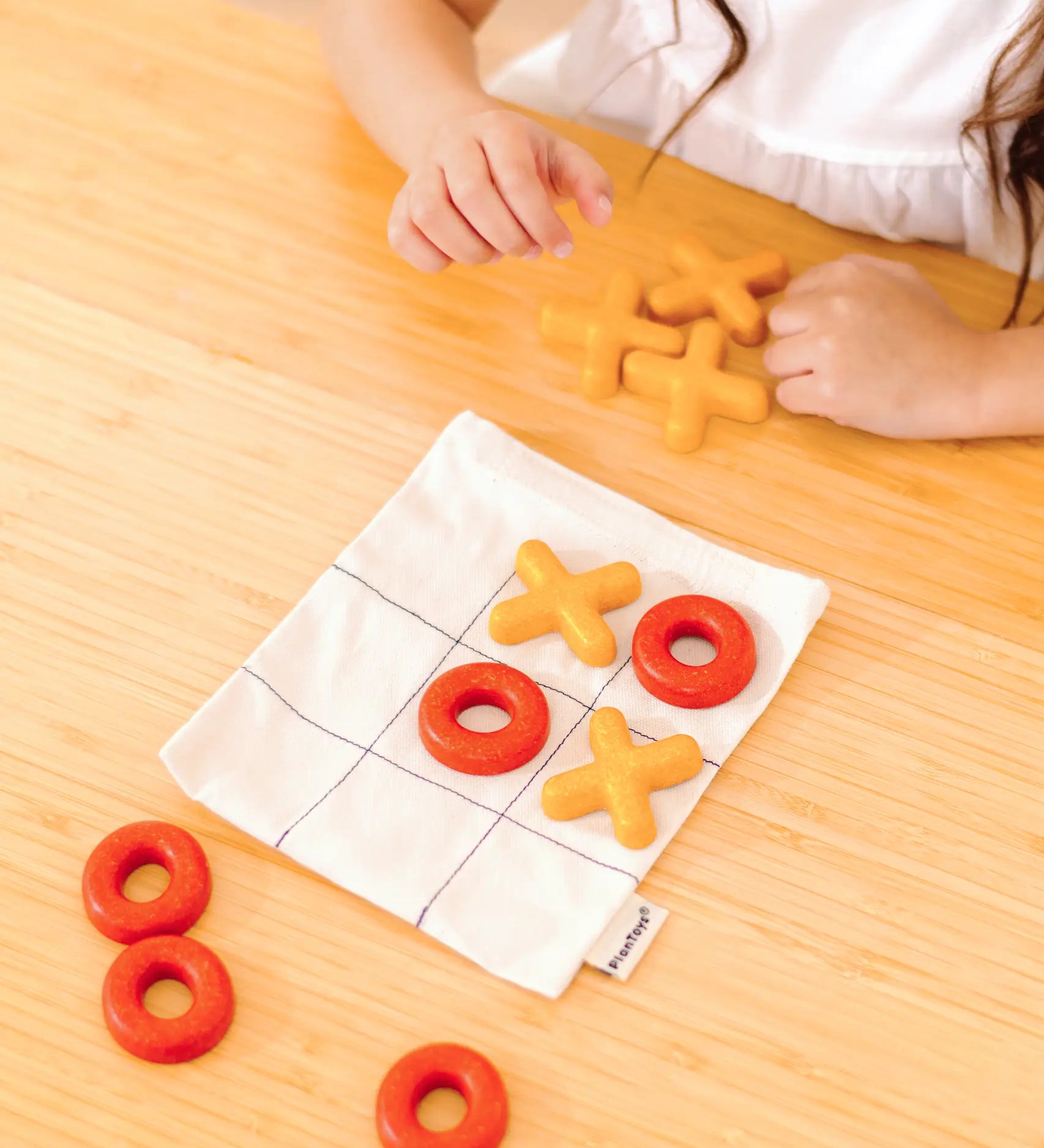 Child playing with the PlanToys Travel Tic-Tac-Toe wooden game set. Cotton bag printed with a grid with 5 yellow x pieces and 5 red o pieces.   