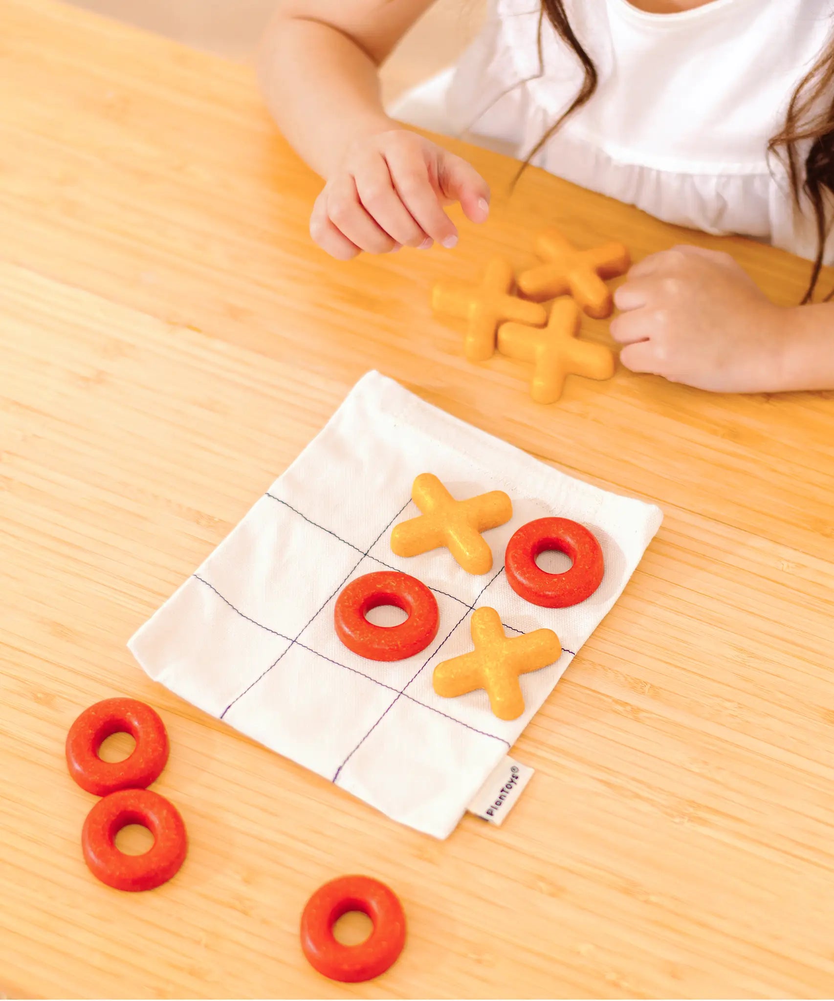 Child playing with the PlanToys Travel Tic-Tac-Toe wooden game set. Cotton bag printed with a grid with 5 yellow x pieces and 5 red o pieces.   