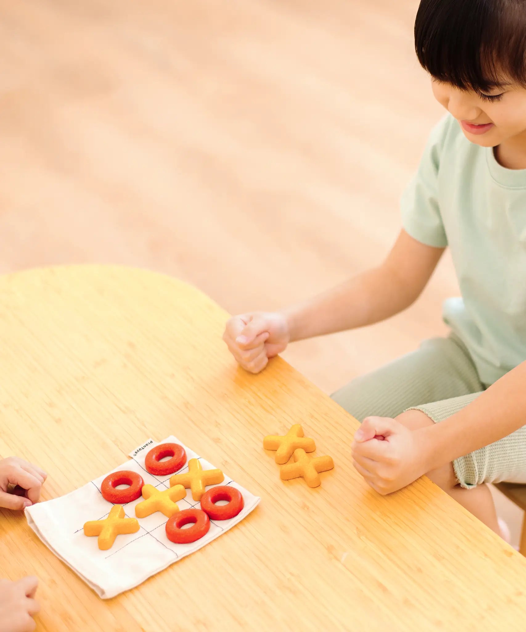 Child playing with the PlanToys Travel Tic-Tac-Toe wooden game set. Cotton bag printed with a grid with 5 yellow x pieces and 5 red o pieces.  