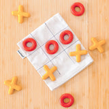 PlanToys Travel Tic-Tac-Toe wooden game set set up on a wooden surface. Cotton storage bag printed with a grid with 5 yellow x pieces and 5 red o pieces.  