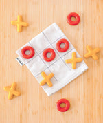 PlanToys Travel Tic-Tac-Toe wooden game set set up on a wooden surface. Cotton storage bag printed with a grid with 5 yellow x pieces and 5 red o pieces.  