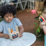 A child sitting my a table playing with the PlanToys Timber Tumble Game, another child is standing near them pretending to take a picture with the PlanToys camera.  