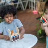 A child sitting my a table playing with the PlanToys Timber Tumble Game, another child is standing near them pretending to take a picture with the PlanToys camera.  