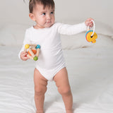 A toddler standing on a bed holding a PlanToys Triangle Clutching Toy in one hand and the key rattle in the other. 
