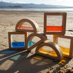 The PlanToys Water Blocks placed on sand on a beach. The light is shining through the blocks casting shadows and colours on the sand. 
