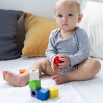A baby sitting on a white blanket playing with the PlanToys kids wooden sensory activity blocks. 