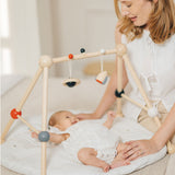 A parent looking at their baby that is laying underneath the PlanToys High Contrast Wooden Baby Play Gym. A wooden baby gym with high contrast coloured features and detailing. The gym comes with two wooden hanging toys, a planet and a rocket. PlanToys range of toys are made from sustainably sourced rubberwood, we're proud stockists of PlanToys here at Babipur. 