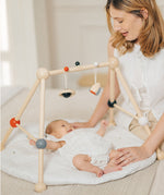 A parent looking at their baby that is laying underneath the PlanToys High Contrast Wooden Baby Play Gym. A wooden baby gym with high contrast coloured features and detailing. The gym comes with two wooden hanging toys, a planet and a rocket. PlanToys range of toys are made from sustainably sourced rubberwood, we're proud stockists of PlanToys here at Babipur. 