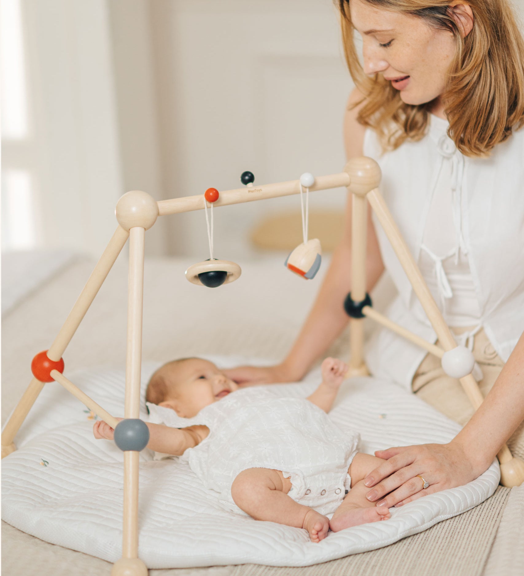 A parent looking at their baby that is laying underneath the PlanToys High Contrast Wooden Baby Play Gym. A wooden baby gym with high contrast coloured features and detailing. The gym comes with two wooden hanging toys, a planet and a rocket. PlanToys range of toys are made from sustainably sourced rubberwood, we're proud stockists of PlanToys here at Babipur. 