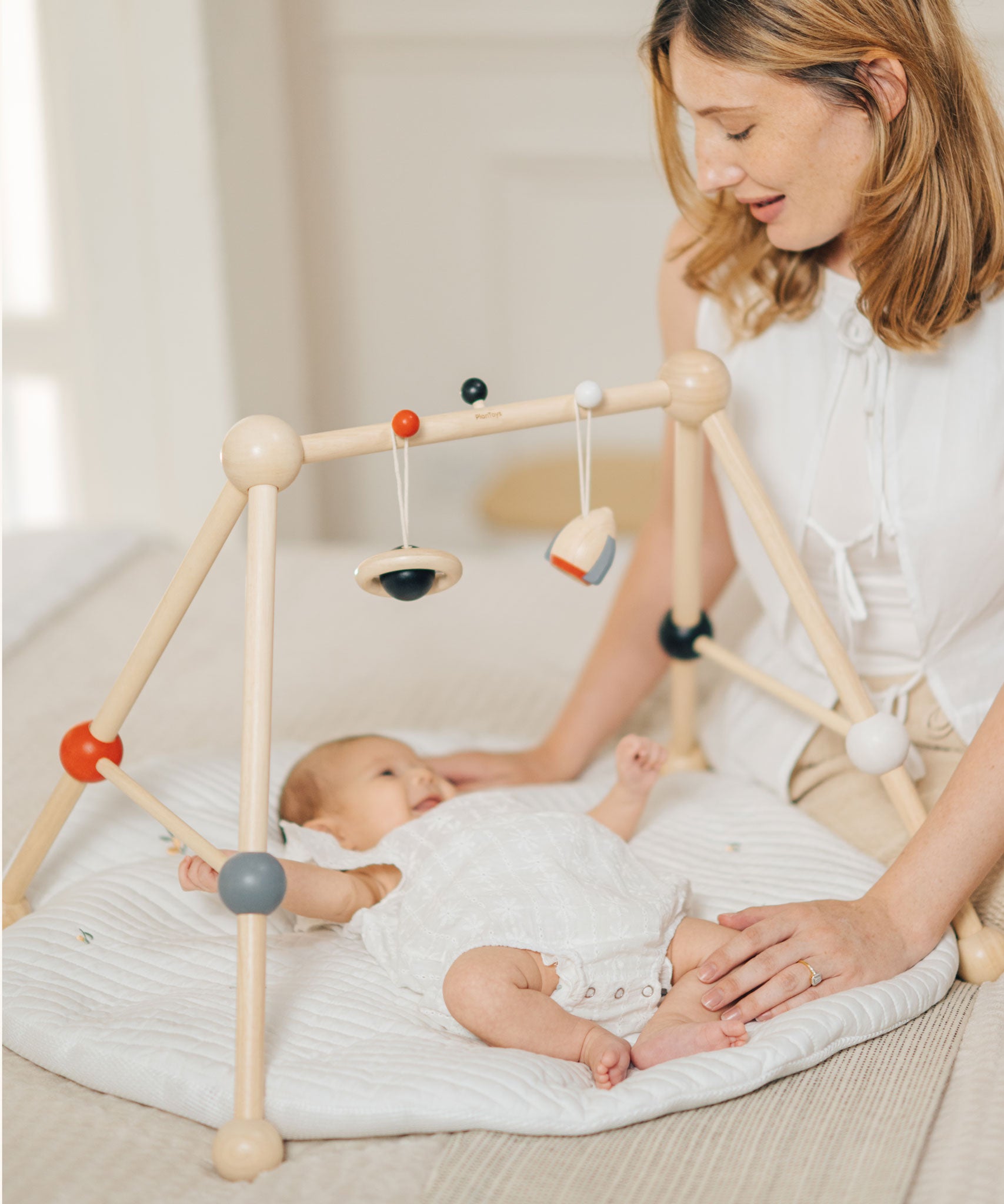 A parent looking at their baby that is laying underneath the PlanToys High Contrast Wooden Baby Play Gym. A wooden baby gym with high contrast coloured features and detailing. The gym comes with two wooden hanging toys, a planet and a rocket. PlanToys range of toys are made from sustainably sourced rubberwood, we're proud stockists of PlanToys here at Babipur. 