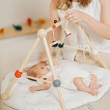 A parent looking at their baby that is laying underneath the PlanToys High Contrast Wooden Baby Play Gym. A wooden baby gym with high contrast coloured features and detailing. The gym comes with two wooden hanging toys, a planet and a rocket. PlanToys range of toys are made from sustainably sourced rubberwood, we're proud stockists of PlanToys here at Babipur. 