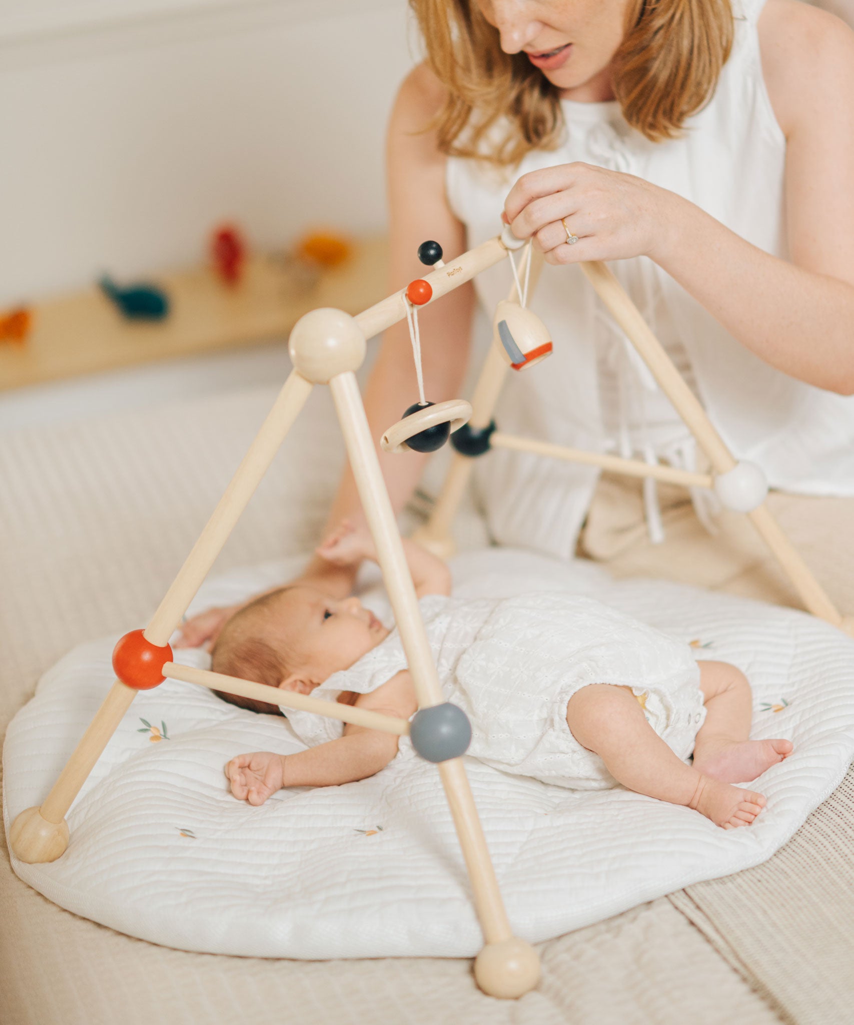 A parent looking at their baby that is laying underneath the PlanToys High Contrast Wooden Baby Play Gym. A wooden baby gym with high contrast coloured features and detailing. The gym comes with two wooden hanging toys, a planet and a rocket. PlanToys range of toys are made from sustainably sourced rubberwood, we're proud stockists of PlanToys here at Babipur. 